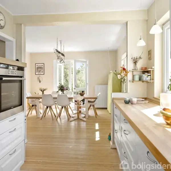 Køkken med hvide skabe og træbordplade, hvor en ovn er integreret. Spisebord med stole står i baggrunden under lysekroner. Rummet er lyst med store vinduer og planter.