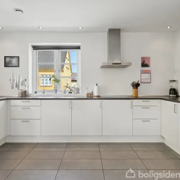 Et moderne køkken med hvide skabe og grå bordplader. Køkkenet har en emhætte, ovne og vindue med udsigt. Væggene er dekoreret med billeder og en plakat.