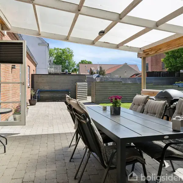 En overdækket terrasse med et sort spisebord og seks stole i en hyggelig gårdhave. Væggene er af mursten, og der er planter på bordet.