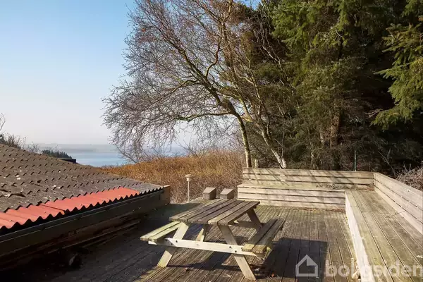 En træbænk og et bord står på en terrasse af træ, omkranset af et hegn. Træer omgiver området, og i baggrunden ses en udsigt over vandet.