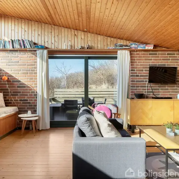 Stue med sofaer placeret foran en stor skydedør, der fører ud til en terrasse. Indretningen inkluderer en indbygget siddeplads, fladskærms-tv, boghylder og planter, omgivet af murstensvægge.