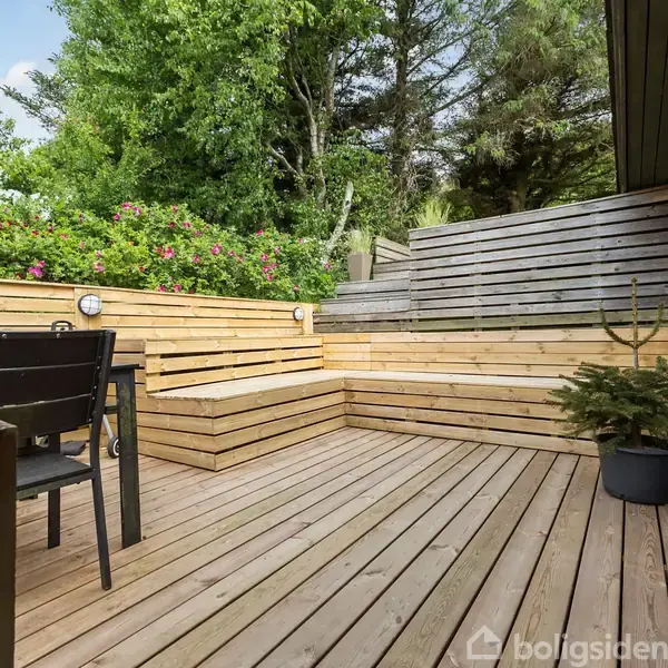 Terrasse i træ med sorte havemøbler og en plante i en potte. Bænke op ad et hegn omgivet af tætte, grønne buske og træer.