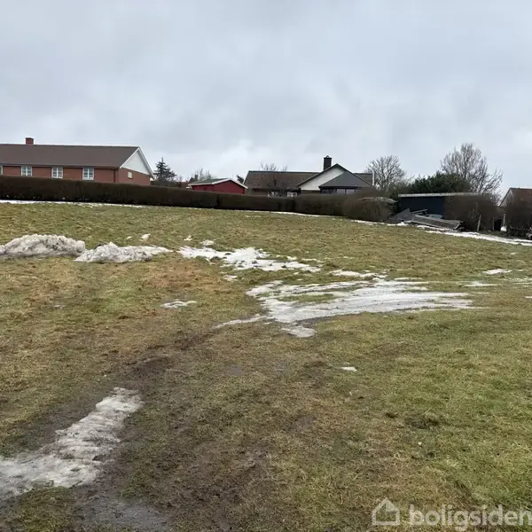 Græsplæne med smeltet sne og spor af køretøjer. Flere huse og en trailer ses i baggrunden under en overskyet himmel.