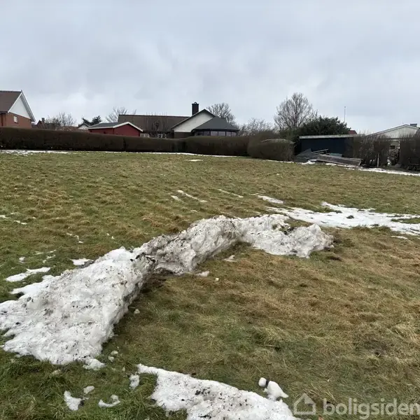 Græsplæne med smeltende snedrive i forgrunden, omgivet af parcelhuse med hække og træer i baggrunden.