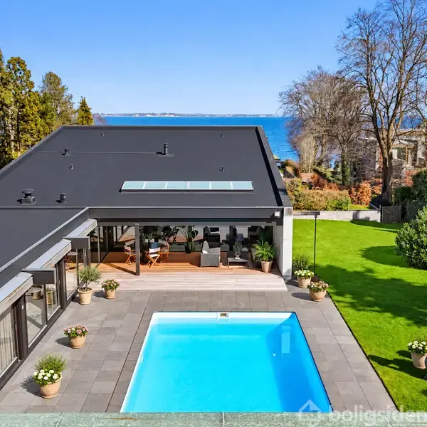 Moderne hus med fladt tag omkranser en blå swimmingpool, omgivet af grønne planter og græsplæne. I baggrunden ses havudsigt og træer, skaber en rolig atmosfære.