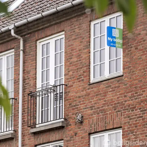 Et rødt murstenshus med hvide vinduer har et til salg-skilt fra "Nybolig" i et af vinduerne. En grøn plante ses i forgrunden.