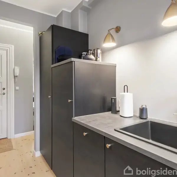 En moderne køkkenvask er placeret i en grå bordplade, omgivet af mørke skabe. To væglamper lyser området. I baggrunden ses en hvid dør og en entre.