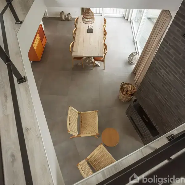 Stol i flet står ved et lille rundt bord i moderne stue. I baggrunden ses et spisebord med stole og store vinduer med gardiner, ved siden af en kurv.