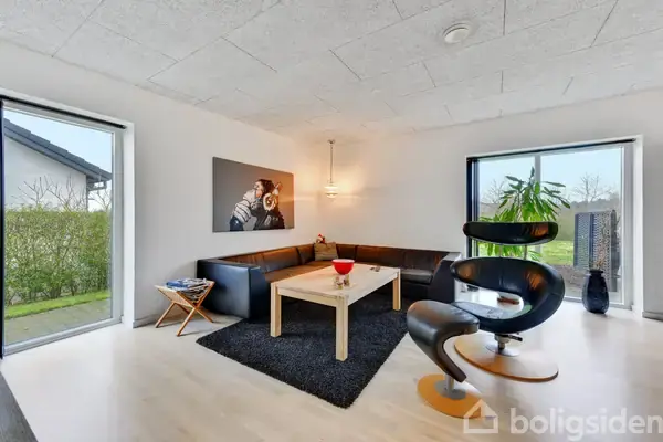 Sort læderstol står på et lyst trægulv, foran en sort lædersofa, i en stue med store vinduer og hvide vægge. Et maleri hænger over sofaen.