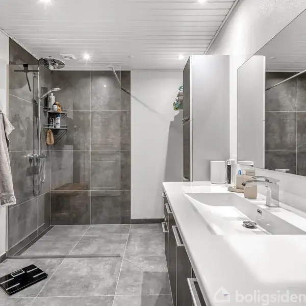 Badeværelse med bruser til venstre, håndklæder på en knage. Til højre er der en lang vask med spejl over, omgivet af hvide fliser og grå gulvfliser.