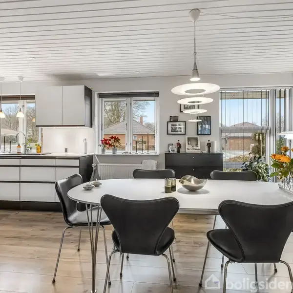 Et moderne køkken med hvide skabe og en sort-hvid mønstret bordplade. Rundt spisebord med sorte stole og en pendel hænger over det. Store vinduer slipper dagslys ind.