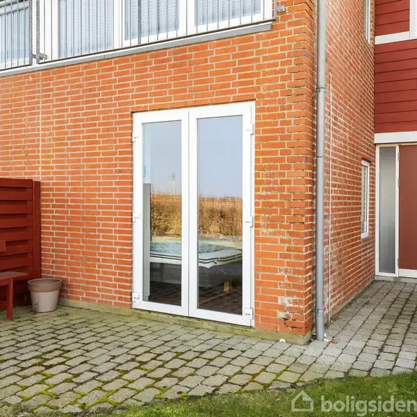 Rødt murstenshus med en dobbelt glasdør, der åbner mod en flisebelagt terrasse. Til venstre en rød træbænk ved siden af en plantekrukke. Husnummer 23 ses ved indgangen.