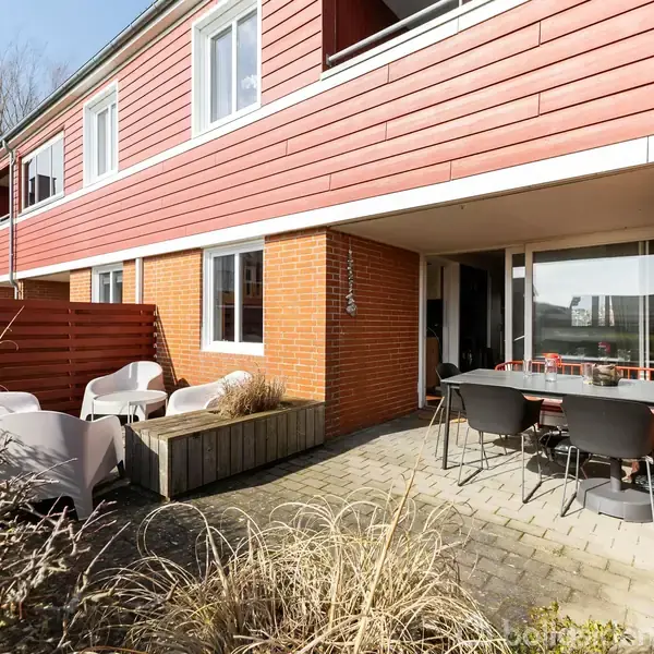 En rækkehusbygning med røde træfacader og store vinduer. På terrassen står udendørsmøbler og en plantekasse. Der er en hæk og et hegn i baggrunden.
