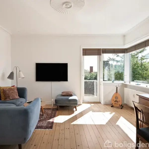 En stue med en blå sofa og fodskammel foran en vægmonteret TV. Ved siden af står en gulvlampe. Et spisebord med stole er nær vinduer med udsigt til grønt træer. En dekorativ vase står ved vinduet.