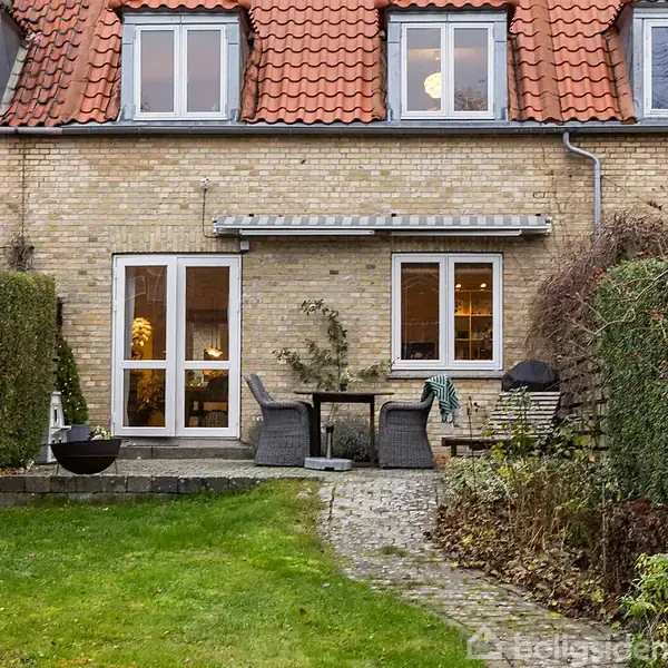 Husfacade med gul mursten og rødt tegltag. Foran er en terrasse med et bord og to stole, flankeret af en buksbomhæk. Til venstre ses havedøre og til højre et vindue.