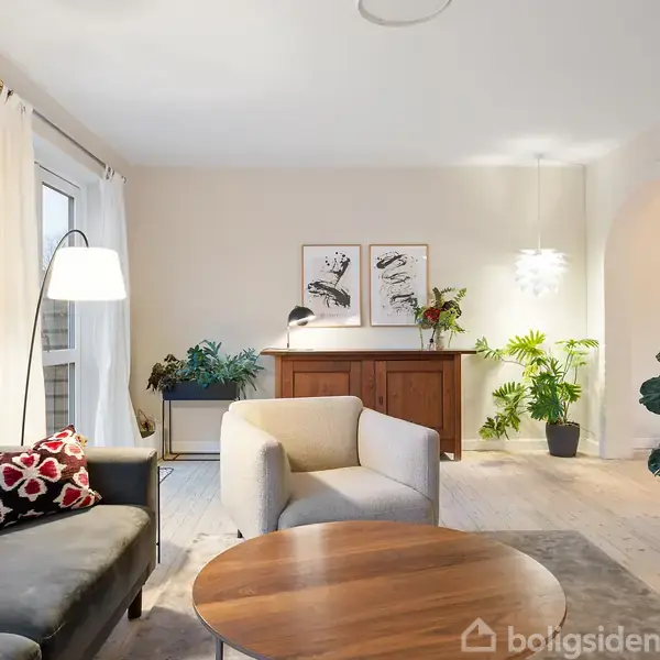 En stue med en sofa og lænestol omkring et rundt sofabord. I baggrunden er en kommode med planter og kunst på væggen. Lyset er blødt og rummet er stilfuldt indrettet.