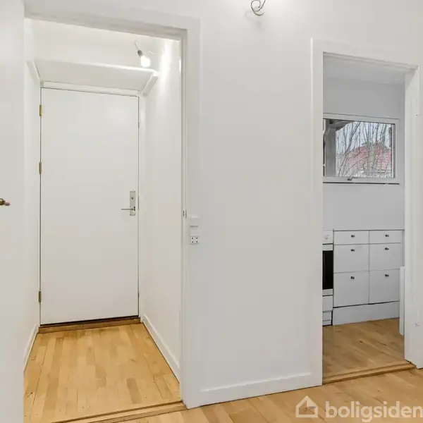 Dør i hvidt rum står lukket, omgivet af lyse vægge og trægulv. Til højre ses en åbning til et køkken med skabe og et vindue.