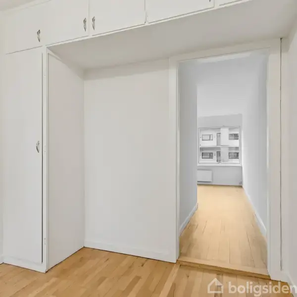 Gang med trægulv fører til en større lys stue med vinduer. Skabe og radiator er placeret i gangen, der har hvide vægge og døre.
