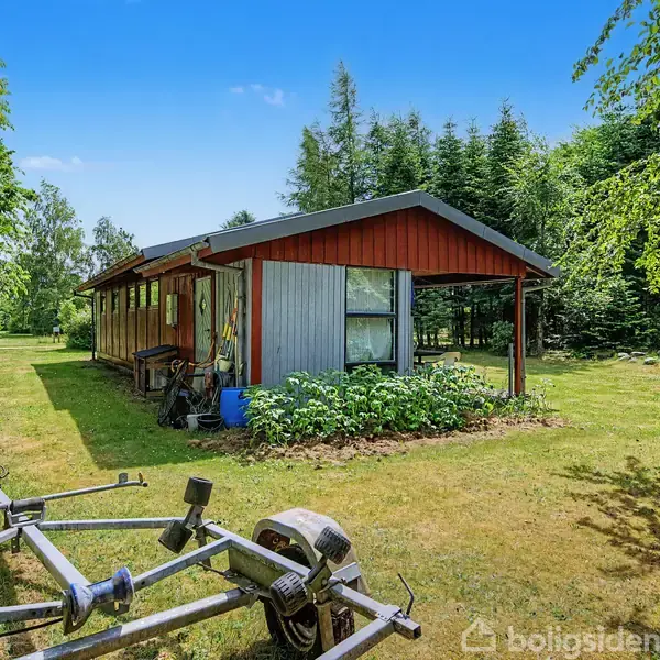 En træbygning med store vinduer står stille i en grøn have omgivet af træer. En bådtrailer ses i forgrunden på græsplænen.