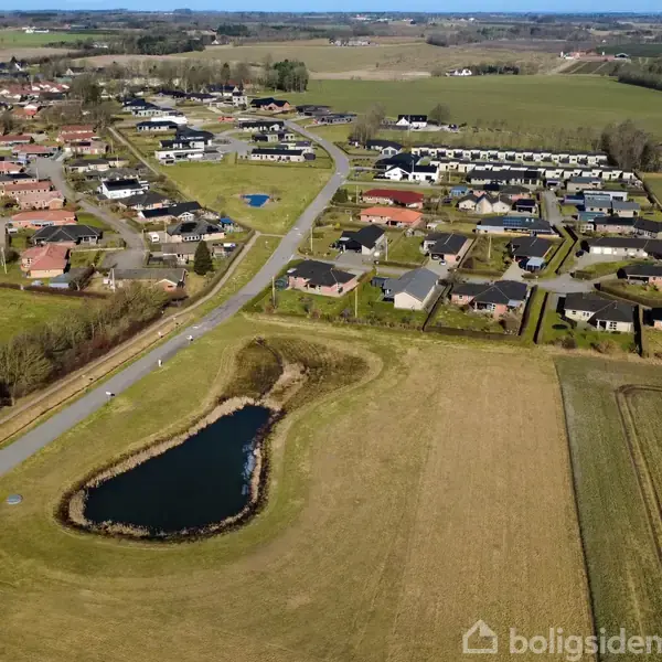 En landsby med en vej gennem grønne marker og et lille dam til venstre. Området er fyldt med huse, og markerne omgiver bebyggelsen.
