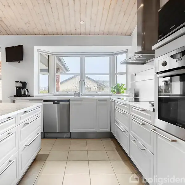 Et moderne køkken med hvide skabe og indbyggede apparater. Rummet har store vinduer, der slipper lys ind, og en kaffeapparat står på bordpladen nær en flaske olie.