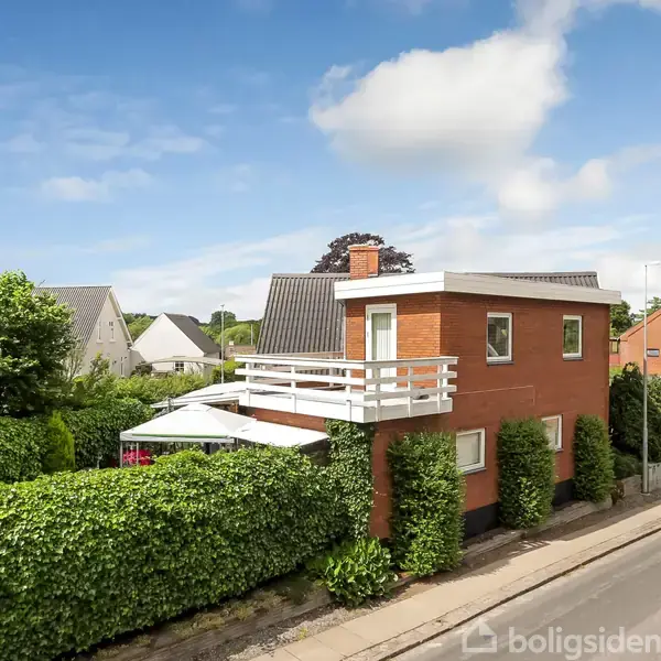 En murstensbygning med hvide altaner står ved en vej omgivet af grønne hække og træer. Flere nabolagshuse er synlige i baggrunden under en blå himmel med få skyer.