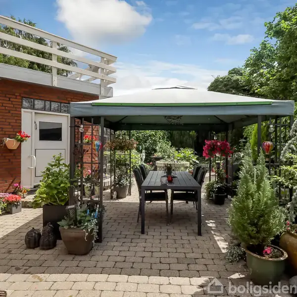 En terrasse med et havebord og stole under en pavillon. Omgivet af krukker med planter, ved siden af en murstensbygning, omgivet af frodigt grønt.