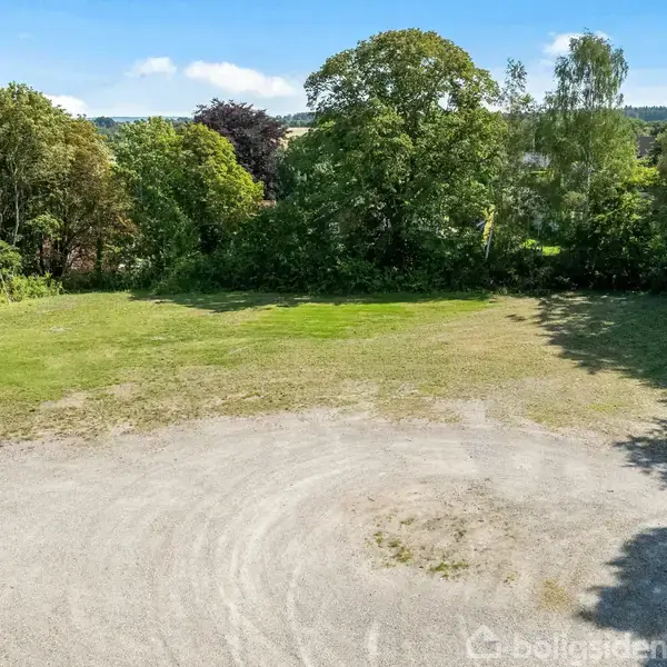 En stor åben grusplads ligger i forgrunden, omkranset af grønne træer og buske under en klar blå himmel uden tekst.