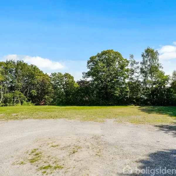 Grusplads foran en græsplæne omgivet af træer under en blå himmel.