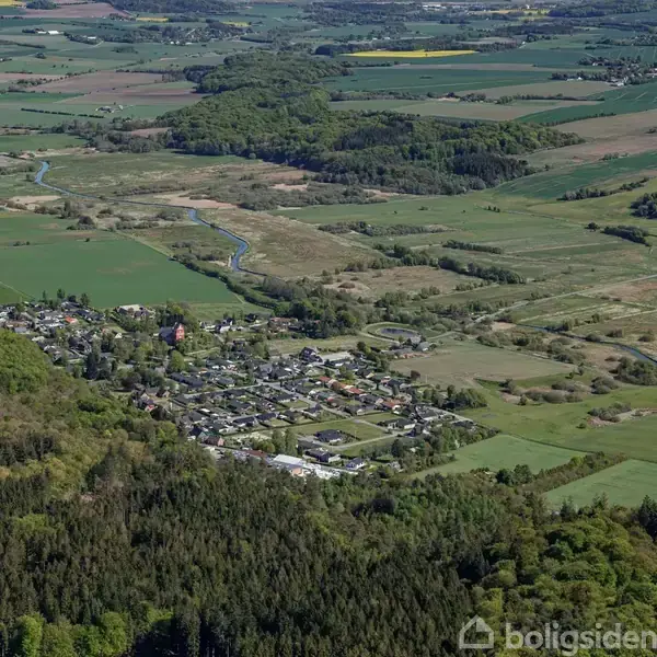 En landsby ligger spredt med huse og bygninger. Den er omgivet af åbne marker og skovområder, set fra oven i et bakket landskab.