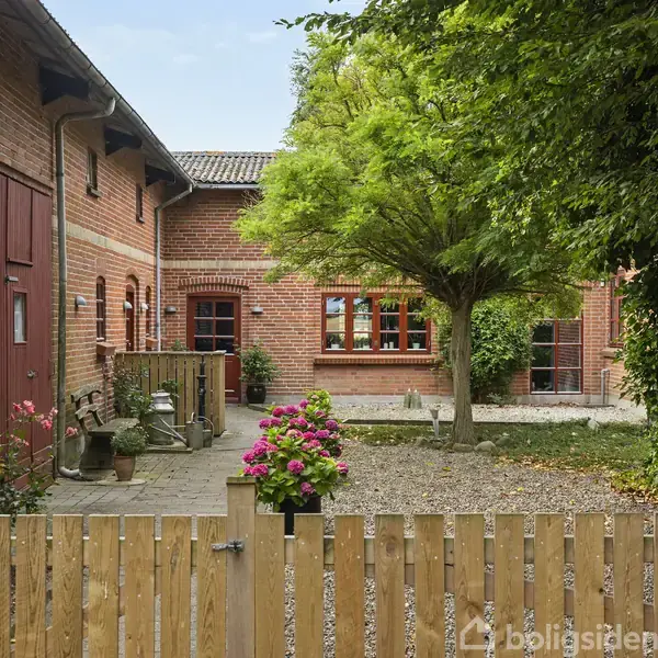 En gårdhave med træhegn foran en rød murstensbygning. En stor busk står ved siden af en flisebelagt terrasse med planter og blomsterpotter. Der ses også en stor træport.