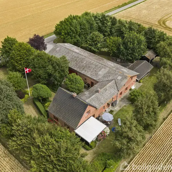 En rød murstensgård omgivet af træer og marker. Et dansk flag vajer i haven, hvor der er græsplæne og nogle blomsterbede. Gården ligger i et landligt område.