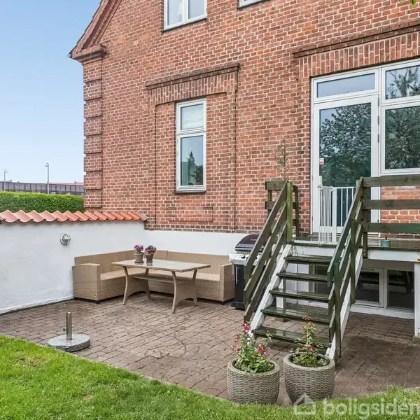 Murstenshus med trappe fører til terrasse med havemøbler og grill. Terrasse omgivet af lav mur, græsplæne og hæk i haveområde.