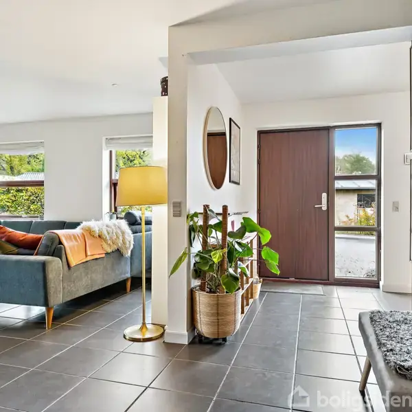 En stue med en blå sofa og flere puder står ved store vinduer, der slipper lys ind. Ved siden af er en dør med glaspaneler. På væggen hænger en række kunstbilleder.