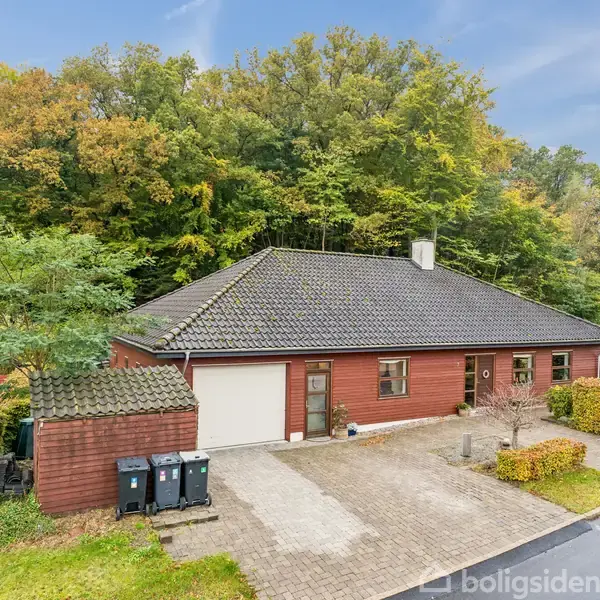 Rødt etplanshus står stille ved en stenbelagt indkørsel, omkranset af grønne hække og høje træer. Skraldespande placeret ved siden af garagen.