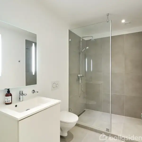Toilet og håndvask med spejl står i et moderne badeværelse. En bruseniche med glasvæg i baggrunden. Rummet er lyst med en minimalistisk indretning. Flasken ved håndvasken mangler tekst.