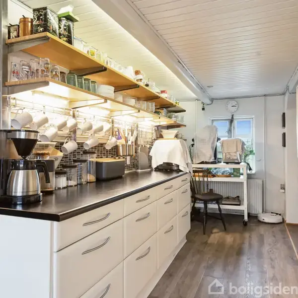 Køkkenbord med kaffemaskine og krukker, hylde med kopper og glas ovenfor. En lys, lang køkkenindretning i en moderne stil med træfarvet gulv, hvide skabe og væghængt mikrobølgeovn.