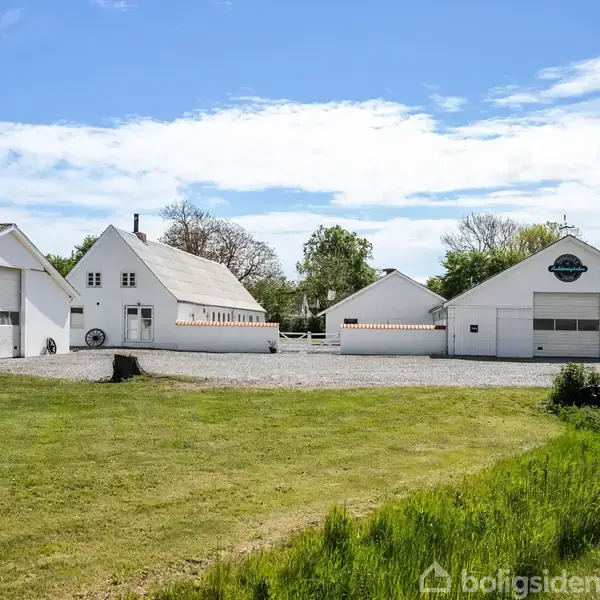 Gårdbygninger med hvide tage og en gårdsplads omgivet af veje og grønne marker. Bagved ses en dam og flere træer, samt spredte huse i det frodige landskab.