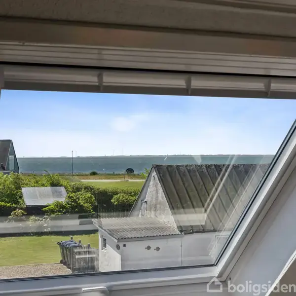 Et skråt tagvindue åbner ud mod en udsigt over en grøn forhave og en dybblå havhorisont i baggrunden. Korte tage og buske er synlige udenfor.