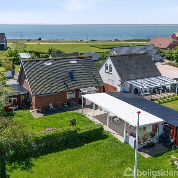 Hus med terrasse og have står stille i et nabolag nær en kystlinje med udsigt over havet. Flere huse og grønne områder omgiver det, skaber en fredfyldt atmosfære.
