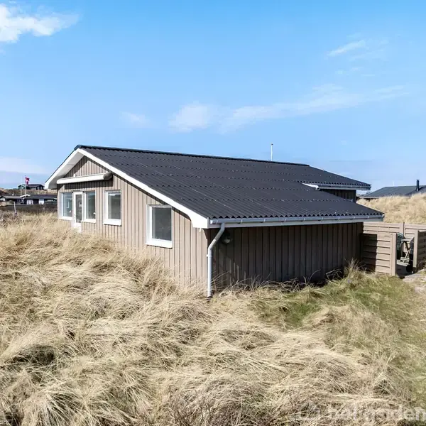 Et træhus står stille på en græsklædt klit, omgivet af tørt græs under en blå himmel. Flere huse er spredt i baggrunden.