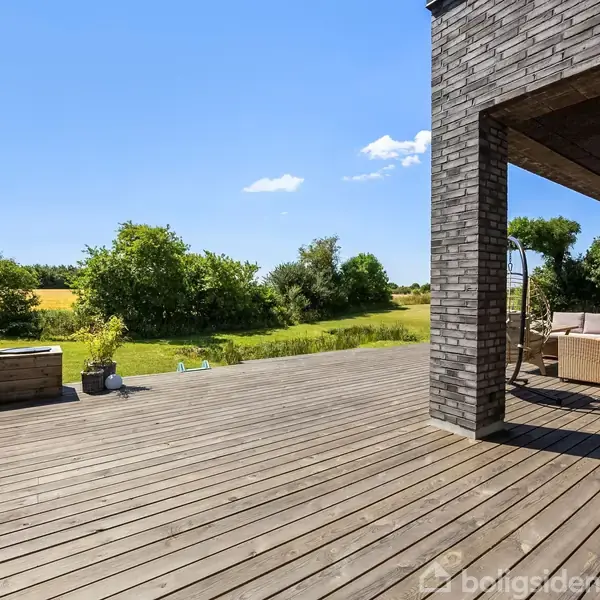 Træterrasse med komfortable sofaer under en overdækket sektion. Omkranset af frodig natur og udsigt over marker under en klar blå himmel.