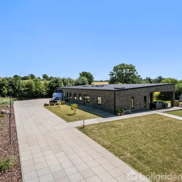 En rektangulær, moderne étplans murstensbygning står stille i et åbent område med en velplejet græsplæne. Flisebelagt indkørsel leder op til huset, omgivet af buske og marker.