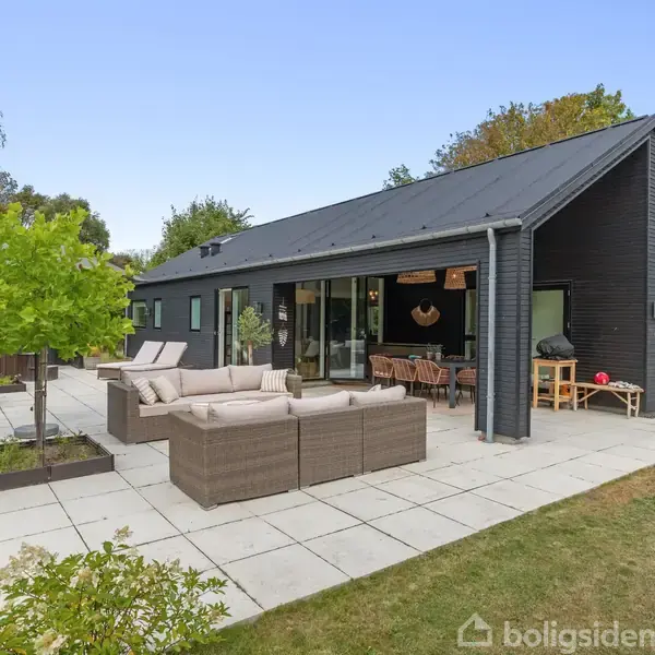 Et moderne, sort træhus med store vinduer har en indrettet terrasse i forgrunden. Terrassen har grå fliser, loungemøbler og grønne planter. Omkringliggende have med træer og buske.
