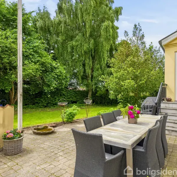 Et havebord med sorte stole står på en terrasse med brosten. Blomster på bordet. Trapper fører op til et gult hus. Baggrund med tæt grøn vegetation og en græsplæne.