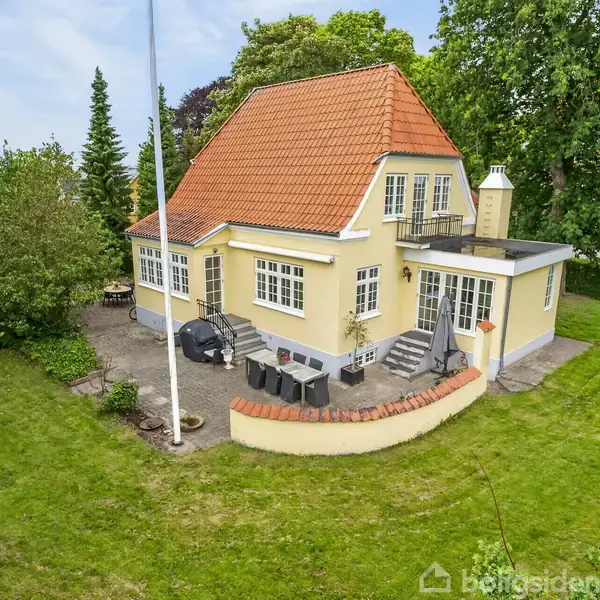 Gult hus med rødt tegltag står i en grøn have. Der er havemøbler på terrassen og en flagstang. Omgivet af træer og buske giver en fredelig atmosfære.