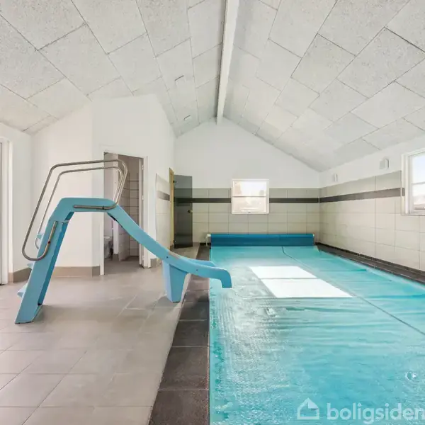 Blå rutchebane fører ned i en indendørs swimmingpool. Poolen befinder sig i et lyst rum med skråt loft og flisegulv. Store vinduer og en glasdør viser en terrasse udenfor.