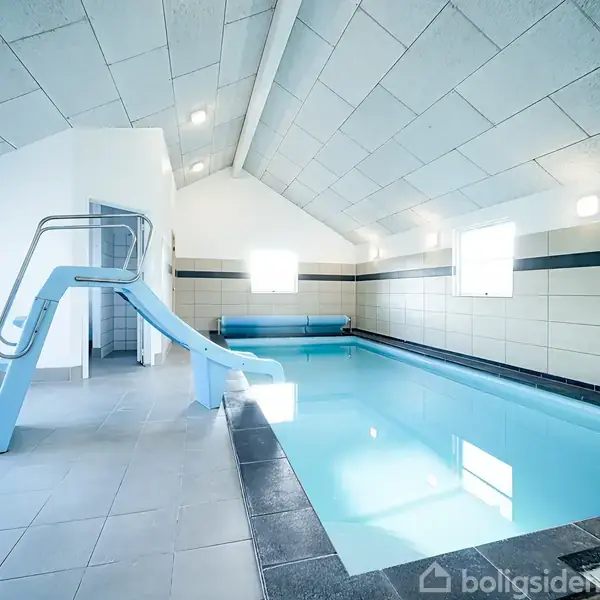 Indendørs swimmingpool med en lyseblå rutchebane, der strækker sig ind i vandet. Omgivet af lyse fliser under et skråt, hvidt loft, med store vinduer der lukker dagslys ind.