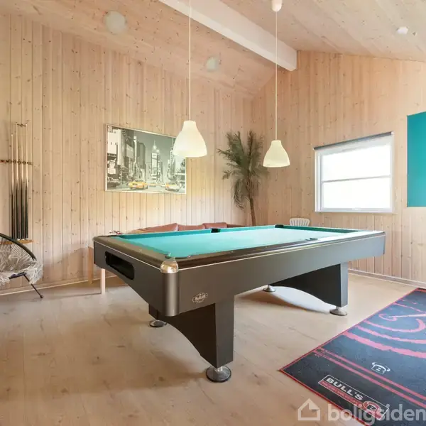Poolbord står i et lyst træbeklædt rum med en dartskive og plakat på væggen. Der er en stol i hjørnet, og to lamper hænger over bordet.