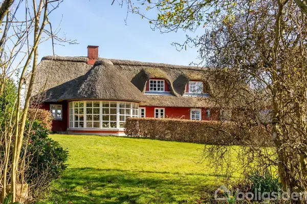 Stråtækt bindingsværkshus i rød farve står stille omgivet af en velplejet græsplæne og buske. Ejendommen har en halvcirkelformet vinterhave og ligger i et fredeligt, frodigt landskab.